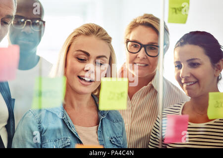 Smiling group of young businesspeople divers remue-méninges avec les notes sur un mur de verre tout en se tenant dans un bureau moderne Banque D'Images