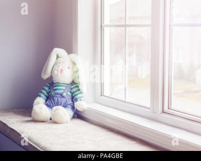 Cute adorable vieux soft plush toy lapin avec de grandes oreilles assis sur windsill près de fenêtre, tonique avec des filtres Instagram, ancienne rétro vintage film Banque D'Images