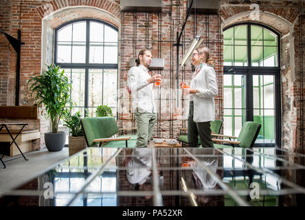 Jeune couple vêtu de blanc se tenant ensemble avec boissons pendant la conversation dans la magnifique loft spacieux intérieur Banque D'Images