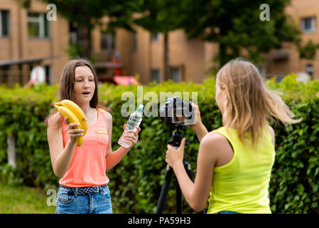 Les jeunes filles des blogueurs, à l'été dans la ville à la nature. Enregistrer une vidéo sur l'appareil photo. Dans ses mains tient une bouteille d'eau et deux bananes. Le conce Banque D'Images