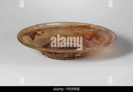 Plat en verre. Culture : Roman. Dimensions : Hauteur : 1 1/4 in. (3.2 cm) Diam. : 6 1/4 x 3 1/2 in. (15,9 x 8,9 cm). Date : 4ème siècle de notre ère. Avec teinte incolore jaune. Le torchage, tubulaire, rim dépliées, bas, et dans, avec d'étroites, légèrement convexe épaulement interne ; S-côté en forme de corps, virant à l'integral en anneau de base tubulaire ; fond concave, épaissie au centre et des traces de petits pontil marque. Intact ; bulles ; piqûre de corrosion, ternissement et irisés, avec la plupart des protections extérieures couvertes en émail épais-comme l'altération. Musée : Metropolitan Museum of Art, New York, USA. Banque D'Images