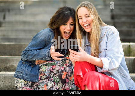 Deux jeunes femmes à la recherche à un drôle de chose sur leur téléphone intelligent à l'extérieur, assis sur les marches. Les filles d'amis à rire. Banque D'Images