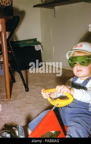 Garçon de deux ans portant des salopettes en velours côtelé, des friandises et un casque au volant d'une voiture de course en peluche, aux États-Unis dans les années 1960 Banque D'Images