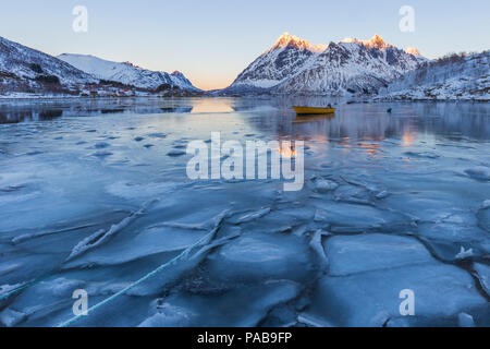 Scène d'hiver de bateau dans le fjord partiellement gelé et paysage enneigé. La glace et l'eau bleue avec des reflets de sommets de montagnes en orange au coucher du soleil. Banque D'Images