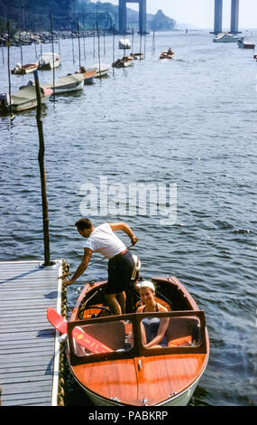 Un couple part en bateau à moteur depuis un quai avec des skis nautiques. Une jeune femme porte un maillot de bain et un homme se déporte de la jetée. Hudson River, Tarrytown, État de New York, États-Unis en été avec des jambes de Tappan Zee Bridge visibles dans les années 1950 Banque D'Images