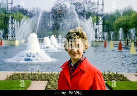 Belle femme portant un manteau rouge souriant devant des fontaines et une porte élaborée dans Pleasure Gardens, Festival Gardens, Battersea Park, Borough of Wandsworth, Londres, Royaume-Uni dans les années 1960 Banque D'Images