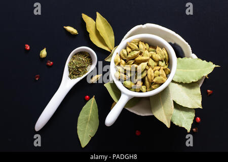 Concept alimentaire à base de plantes vertes Mélange des épices biologiques les feuilles de laurier, poivre rose et gousses cadamom dans maïs tasse en céramique blanche sur fond noir Banque D'Images