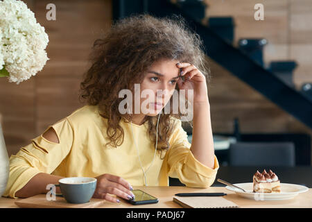 Déprimé jeune femme avec des cheveux bouclés assis sur la table du restaurant en bois, épuisé et en colère, à sentir les émotions négatives. Tasse de café et fleurs lilas blanc, escaliers sur arrière-plan. Banque D'Images