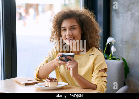 Curly girl avec maintien de sourire magnifique tasse de café, en regardant la caméra. Le port de blouse jaune avec des trous sur les manches. Délicieux délicieux morceau de gâteau debout sur une table en bois près de la fenêtre. Style de vie. Banque D'Images