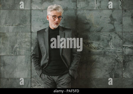 Portrait of handsome man en tailleur debout sur un mur gris. Classy businessman standing with hands in pocket et regardant la caméra. Banque D'Images