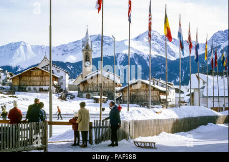 Station de ski autrichienne avec église catholique Pfarramt et des gens qui regardent des patineurs sur une patinoire en hiver et un homme avec un traîneau en bois, Serfaus, Autriche dans les années 1960 Banque D'Images