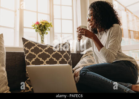 Businesswoman working on laptop computer assis à la maison tenant une tasse de café dans la main. Smiling woman sitting on sofa at home et à la sortir de la wi Banque D'Images