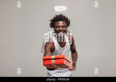 Concept d'amour et de partage. goft barbu teint foncé Angel de l'Afrique de l'homme drôle avec chemise blanche, nimbus et des clins d'atanding et holding present gift box. Banque D'Images