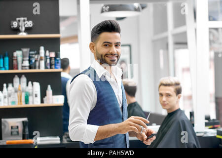 Smiling coiffure debout près de client assis près de miroir, looking at camera. Le port casual chemise blanche, gilet gris élégant. Posant devant des étagères avec les produits de soins capillaires. Un sentiment positif. Banque D'Images