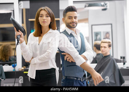 Deux hairstylers posant debout dans le salon moderne, jeune beaty client assis derrière. Parrucchieri portant des chemises blanches classiques, l'homme ayant gilet gris. Maintien de la femme d'un sèche-cheveux, looking at camera. Banque D'Images