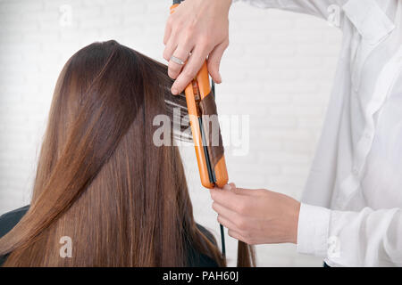 Hairstylist Utilisation de client, en gardant les cheveux bruns, ce lisseur curles. Modèle avoir long et sombre, droit, lisse, glancy cheveux. Coiffure portant chemise blanche classique. fond blanc. Banque D'Images