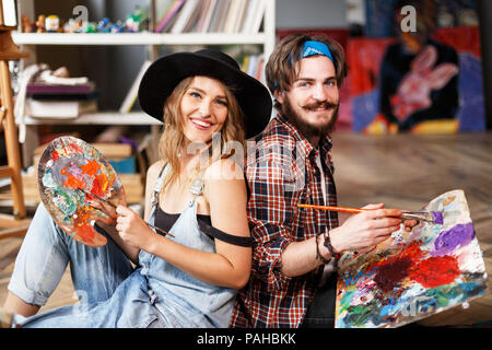 Deux artistes pretty smiling habillés en vêtements hippie holding pinceaux et couleurs mélangées sur la palette en studio contemporain avec de nombreuses peintures Banque D'Images