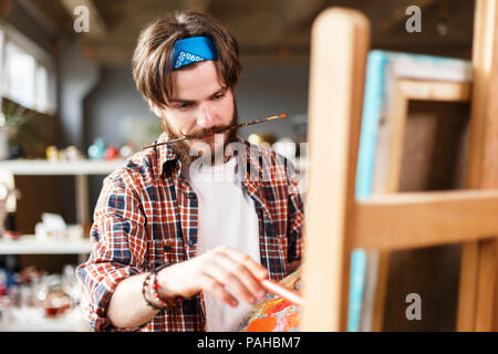 Dark-haired créative hipster barbu artistes masculins en bleu bandana holding palette et encombrement derrière le chevalet dans son studio contemporain avec de nombreux tableaux, tenant un pinceau dans ses dents Banque D'Images