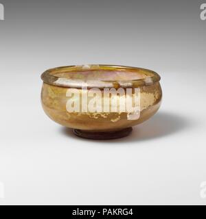 Bol en verre. Culture : Roman. Dimensions : Hauteur : 1 7/16 in. (3,7 cm) Diam. : 3 1/4 x 1 1/2 in. (8,3 x 3,8 cm). Date : 2ème-3ème siècle de notre ère. Jaune verdâtre translucide. Jante pliée vers l'extérieur et vers le bas, formant en partie arrondie, col tubulaire à la bouche à lèvre intérieure ; côtés bombés en courbe à l'integral, faible anneau de base tubulaire ; fond plat, avec des traces de pontil et un léger coup de pied. Intacts ; peu de bulles ; ternissement, de légères piqûres et iridescence sur l'extérieur, crémeux, altération irisé sur l'intérieur. Musée : Metropolitan Museum of Art, New York, USA. Banque D'Images