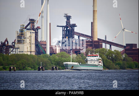 Cargo navigant sur une rivière avec feuillage vert, immense aciérie et plusieurs éoliennes dans l'arrière-plan Banque D'Images