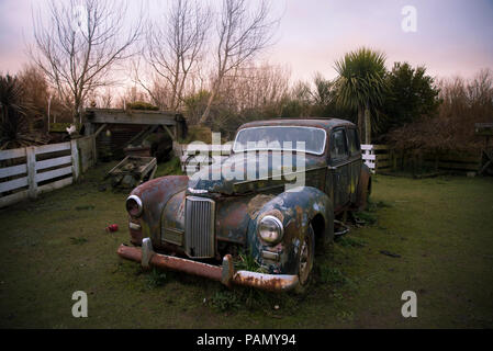 Old rusty vintage voiture Humber en plus d'une clôture blanche, sur l'herbe dans un jardin, au coucher du soleil. Banque D'Images