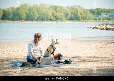 Femme assise sur couverture à une rivière avec dog wearing headphones and using laptop Banque D'Images