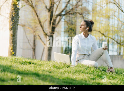 Young businesswoman en pause, à l'aide d'un ordinateur portable, assis dans l'herbe et boire du café Banque D'Images