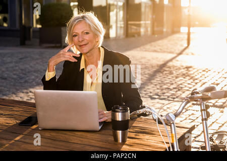 Smiling senior businesswoman using laptop in la ville au coucher du soleil Banque D'Images