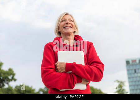 Happy senior woman wearing red hoodie holding laptop outdoors Banque D'Images