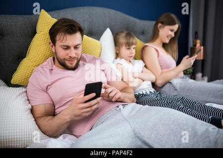 Négligé petite fille assise sur le lit avec ses parents, using smartphones Banque D'Images