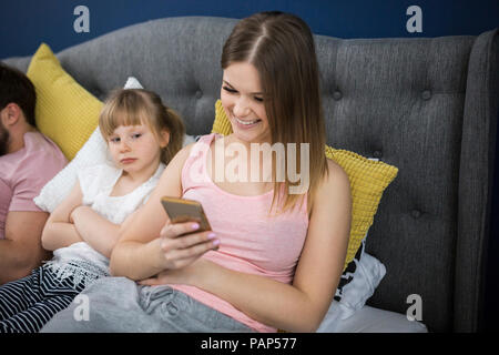 Négligé petite fille assise sur le lit avec ses parents, using smartphones Banque D'Images