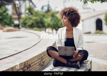 Jeune femme à la mode avec des cheveux bouclés assis sur un banc avec coffre Banque D'Images