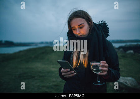 France, Bretagne, Landeda, young woman with cell phone et verre de vin à la côte au crépuscule Banque D'Images