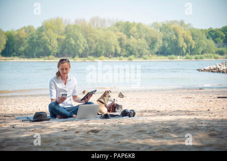 Femme assise sur couverture à une rivière avec chien à l'aide des périphériques portables Banque D'Images