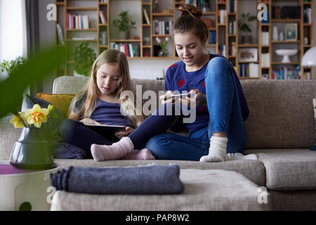 Deux soeurs assis sur le canapé, jouant et catting avec les appareils numériques Banque D'Images