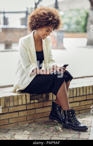 Jeune femme avec des cheveux bouclés sitting on wall looking at cell phone Banque D'Images