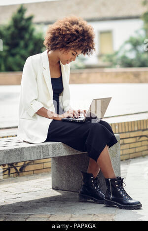 Jeune femme à la mode avec des cheveux bouclés sitting on bench using laptop Banque D'Images