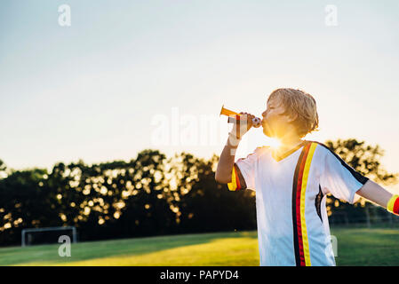 Boy wearing German soccer shirt, soufflant cornes sur terrain de football Banque D'Images