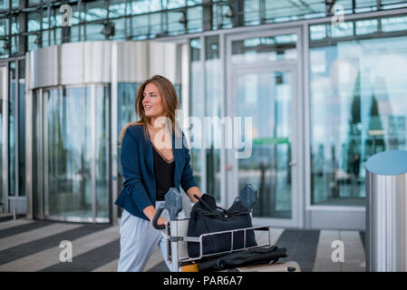 Souriante jeune femme poussant chariot à bagages à la recherche autour de Banque D'Images