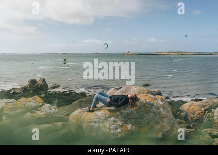 France, Bretagne, Landeda, young woman wearing headphones lying on rock at the coast Banque D'Images