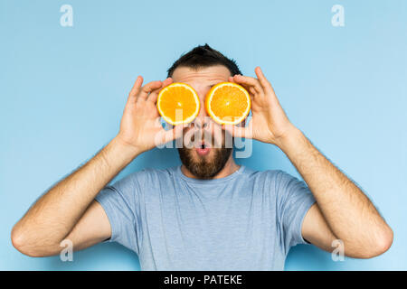 Jeune homme barbu tenant tranches d'orange en face de ses yeux. Fond bleu clair, expression surprise sur le visage de l'homme. Banque D'Images