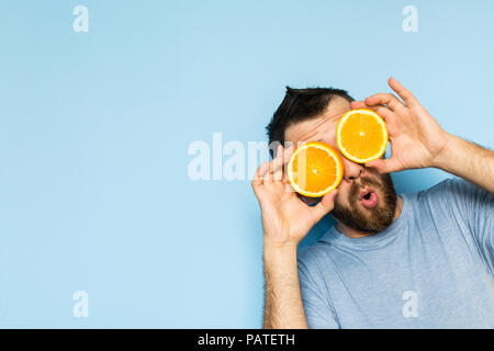 Jeune homme barbu tenant tranches d'orange en face de ses yeux. Fond bleu clair, expression surprise sur le visage de l'homme. Banque D'Images