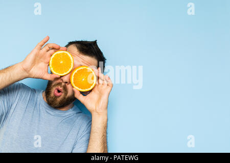 Jeune homme barbu tenant tranches d'orange en face de ses yeux. Fond bleu clair, expression surprise sur le visage de l'homme. Banque D'Images