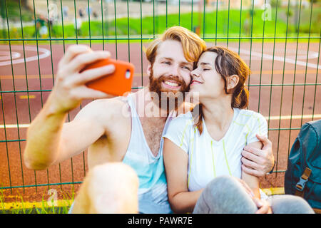 Une paire mans cheveux et la barbe rouge et la femme reste après le sport à l'extérieur sur l'herbe en arrière-plan du stade. Le mec est titulaire d outstre sur smartphone rouge Banque D'Images