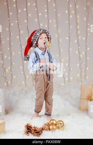 Portrait of white Caucasian boy wearing Santa Claus hat célébrer Noël ou Nouvel An. Adorable petit enfant mignon manger des bonbons en léchant sucette Banque D'Images