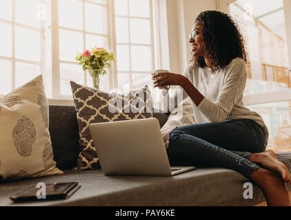 Businesswoman working on laptop computer assis à la maison tenant une tasse de café dans la main. Smiling woman sitting on sofa at home et à la sortir de la wi Banque D'Images