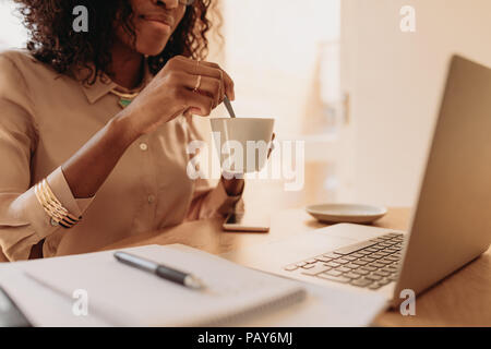 Businesswoman working on laptop computer assis à la maison tenant une tasse de café dans la main. Femme entrepreneur en remuant son café avec cuillère tout en travaillant Banque D'Images