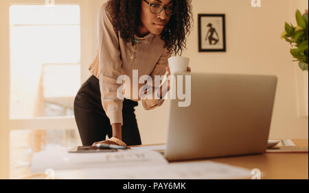 Businesswoman looking at laptop computer tenant une tasse de café dans la main. Femme entrepreneur travaillant sur l'ordinateur portable de la maison debout à côté de la table. Banque D'Images