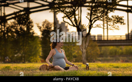 Belle jeune femme enceinte avec petit chien assis sur le tapis, bénéficiant d'yoga, se détendre, se sentir en vie, de respirer l'air frais, de calme et de rêve, dans un parc d'été. sur l'arrière-plan le pont de chemin de fer et de la ville Banque D'Images