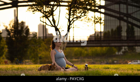 Belle jeune femme enceinte avec petit chien assis sur le tapis, bénéficiant d'yoga, se détendre, se sentir en vie, de respirer l'air frais, de calme et de rêve, dans un parc d'été. sur l'arrière-plan le pont de chemin de fer et de la ville Banque D'Images